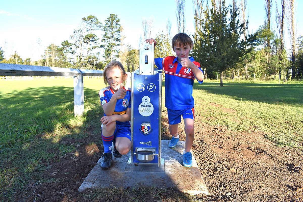 Two boys in soccer uniform standing next to water refill station on sunny day